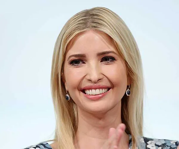 A person with long blonde hair and earrings smiling, clapping hands, against a plain background.