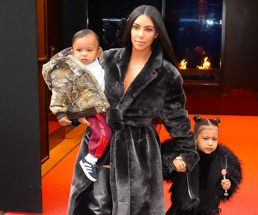 Woman in a fur coat holding a baby and a child with a lollipop, standing on a red carpet.