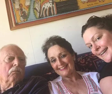 Three people sitting on a couch, smiling, with colorful artwork on the wall behind them.