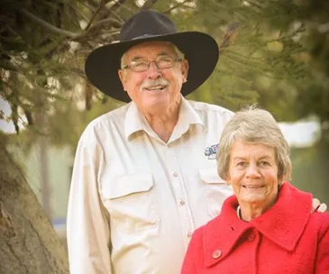 Elderly couple outdoors; man in a cowboy hat and woman in a red coat, smiling under a tree.