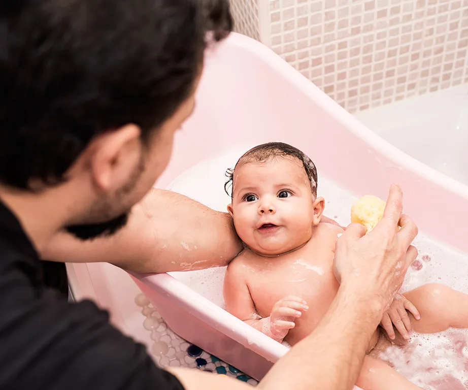 Father bathing infant in a pink baby tub with a sponge, surrounded by bubbles.