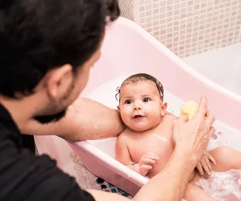 Father bathing infant in a pink baby tub with a sponge, surrounded by bubbles.