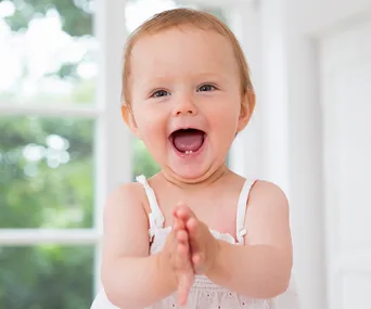 Smiling baby in a white outfit clapping hands in a bright room.