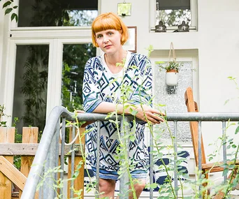 A woman with short orange hair stands on a verandah, leaning on a railing, surrounded by plants.