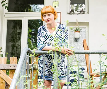 A woman with short orange hair stands on a verandah, leaning on a railing, surrounded by plants.