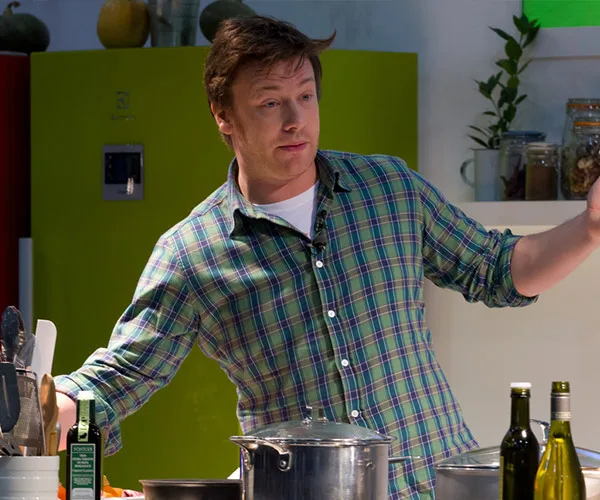 Jamie Oliver cooking in a kitchen, wearing a green plaid shirt, surrounded by pots, olive oil bottles, and utensils.