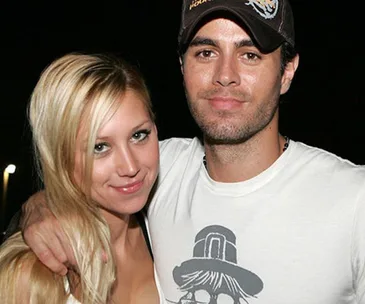 Young woman with long blonde hair smiling, standing closely beside a man in a cap, both in casual attire, at night.