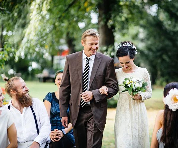 Bride walks down outdoor aisle with man in brown suit, holding bouquet; guests seated, smiling.
