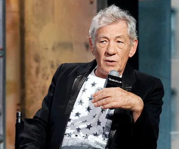 Actor speaking into a microphone during a public event, wearing a star-patterned shirt and blazer.
