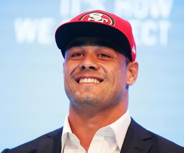 A person smiling, wearing a red cap with a sports team logo, dressed in a suit.