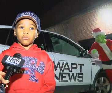 Young boy in a red jacket speaks into a microphone, standing by a WAPT News car with the Grinch dressed as Santa in the background.