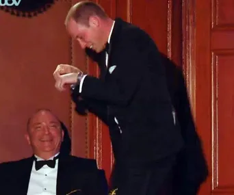 Prince in a tuxedo gallops beside a seated man, entertaining at the Royal Variety Performance.