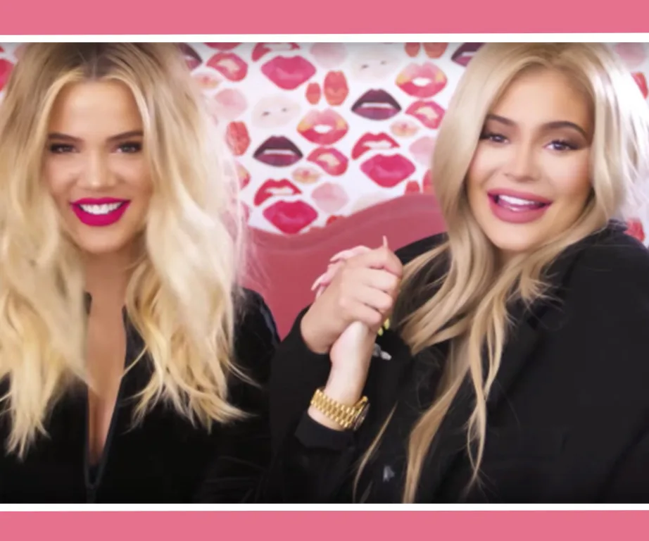 Two women with long blonde hair smiling and holding hands in front of a backdrop with lip prints.