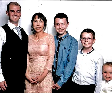 A smiling family portrait with two adults and three children dressed formally, standing against a plain backdrop.