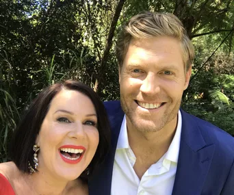 Two people smiling in a lush, green outdoor setting, with one wearing a red top and the other a blue suit.