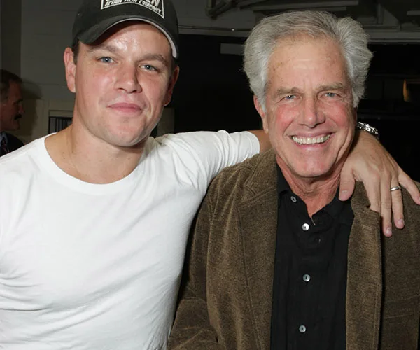 Two men smiling, one in a white shirt and cap, the other in a brown jacket, indoors with arms around each other.