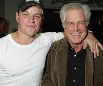 Two men smiling, one in a white shirt and cap, the other in a brown jacket, indoors with arms around each other.