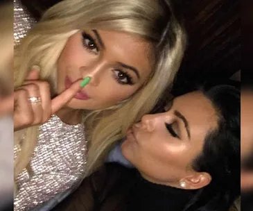 Two women pose closely; one gestures for silence, the other leans in. Both wear glamorous outfits and makeup.