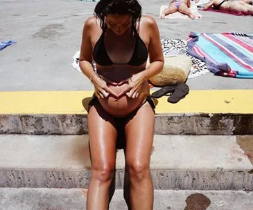 Woman in a bikini sitting on steps, forming a heart shape on her pregnant belly.