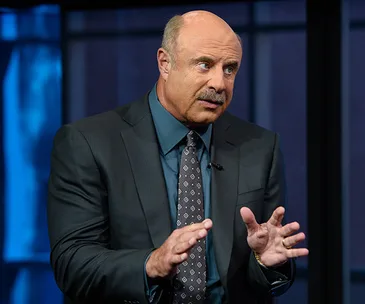 Dr. Phil in a suit gestures while speaking, set against a dark background.