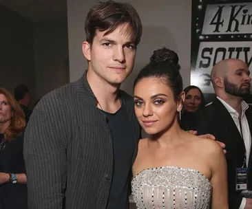 A couple poses together at an event, with a sign and attendees in the background.