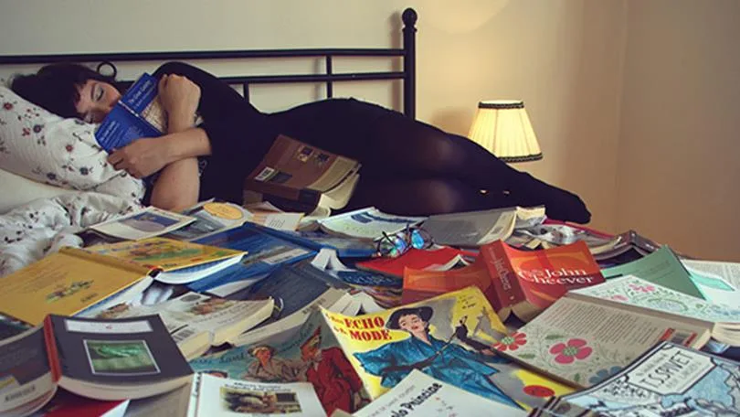 A person sleeping on a bed covered in various open books, with a lamp in the background.