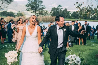 A smiling couple in wedding attire holding hands, surrounded by guests and nature.