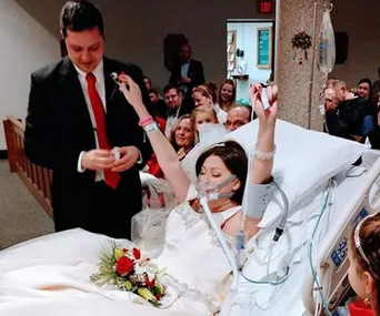 Bride in hospital gown raises arms in joy, lying in a hospital bed, as groom stands by holding wedding ring.