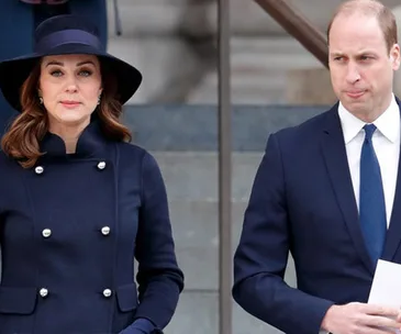 A man and woman in formal attire standing outdoors on steps, woman in navy coat and hat, man in navy suit holding a paper.