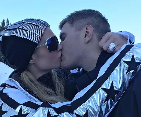A couple wearing winter attire shares a kiss outdoors; a large engagement ring is visible.