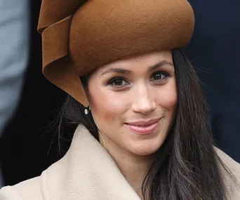 A woman in a brown hat and coat smiles warmly, with long dark hair and earrings visible.