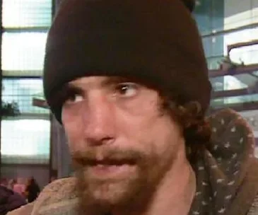 A man with a beard and a beanie is seen in a close-up shot, with an urban background.