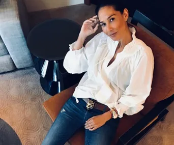 Woman in a white blouse and jeans sitting on a chair, smiling at the camera.