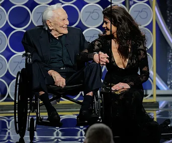 Elderly man in a wheelchair with a woman in elegant attire on stage at a formal event, holding hands and smiling at each other.