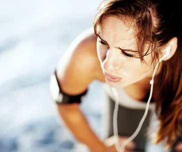 A tired woman in workout gear with earphones, bending over outdoors, suggesting exhaustion.