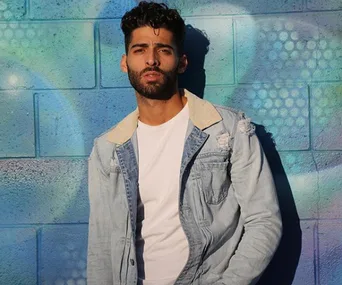Young man in a denim jacket leaning against a colorful graffiti wall, looking confidently at the camera.