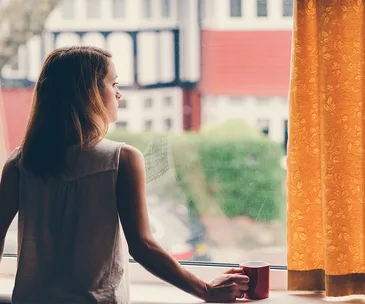 Woman holding a red mug, looking out the window at an urban landscape with red and white buildings.