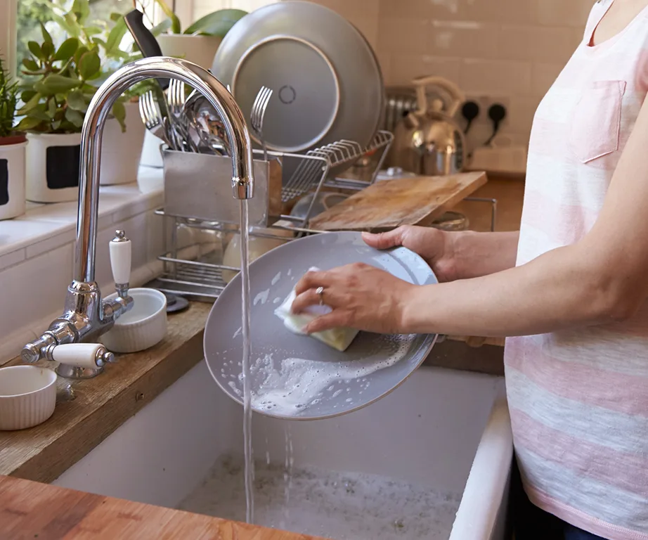 Person washing a plate in a kitchen sink with running water, soap suds visible, and plants in the background.