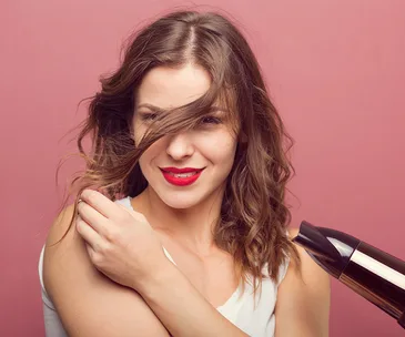 Woman with wavy hair and red lipstick using a blow dryer, smiling against a pink background.