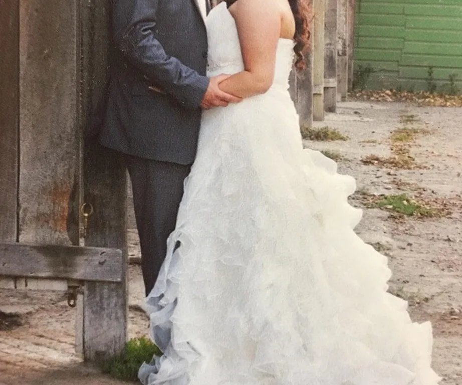 A bride in a ruffled white wedding dress embraces a groom in a dark suit against a rustic wooden backdrop.