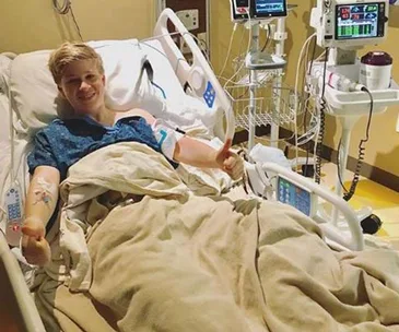Man smiling in hospital bed, giving thumbs up, with medical equipment around.