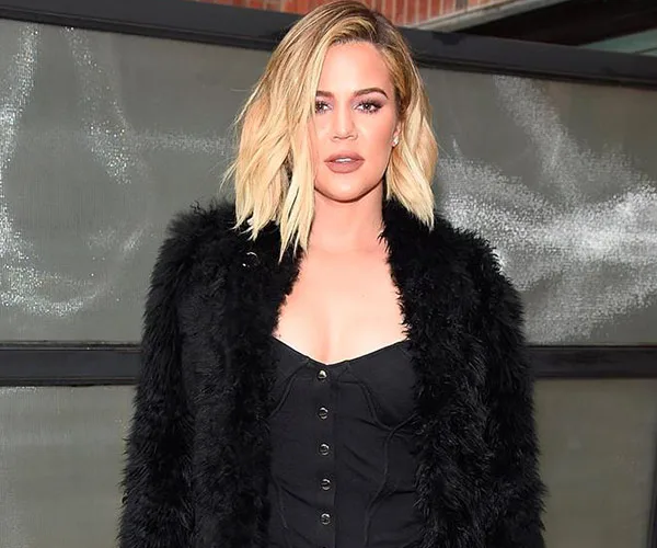 Exterior shot of a woman with blonde hair in a black outfit and fur coat standing in front of a gray backdrop.