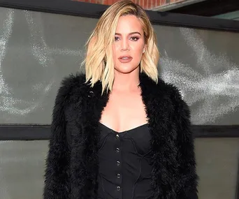 Exterior shot of a woman with blonde hair in a black outfit and fur coat standing in front of a gray backdrop.