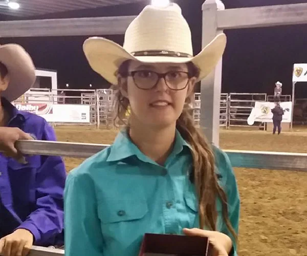 A young person in glasses and a cowboy hat at a rodeo, wearing a turquoise shirt and holding an award.