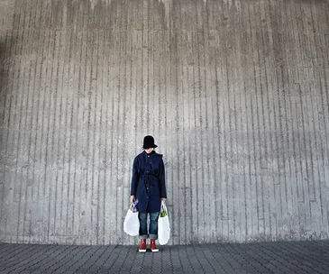 Person in blue coat and hat holding plastic bags stands against large concrete wall, conveying solitude and modern urban life.