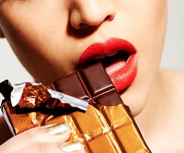 A person with red lipstick eating a partially unwrapped chocolate bar.