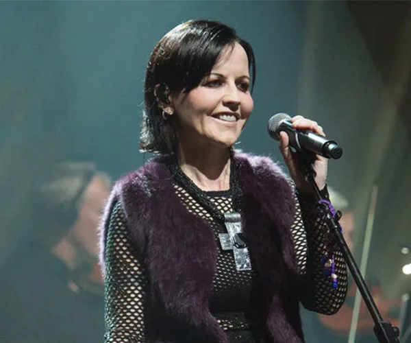 Singer Dolores O'Riordan performing on stage, holding a microphone and smiling.