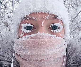 Person bundled up in winter gear with frost on eyelashes, face covered by scarf, in snowy Oymyakon, Russia.