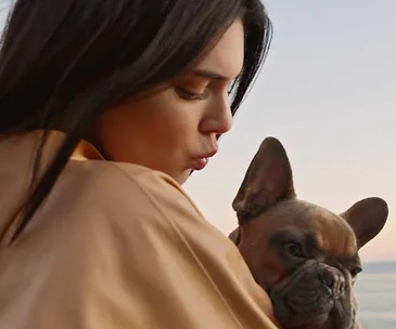 A woman affectionately holds a small dog outside, near the beach, at sunset.