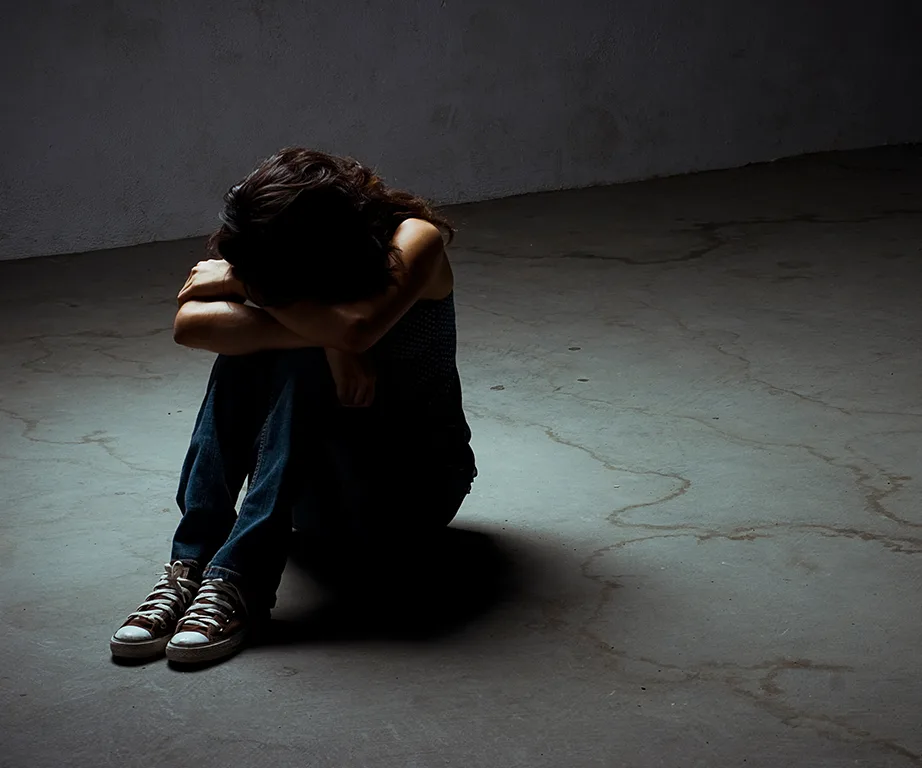 A person sitting on the floor in a dimly lit room, hugging their knees, with a sense of solitude and distress.